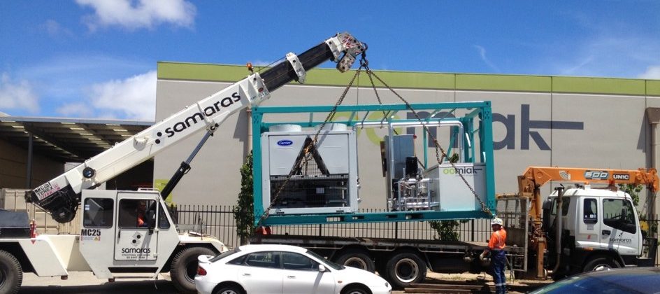 a crane lifting a containerized chilled water supply rig onto a truck with a white car in front of it