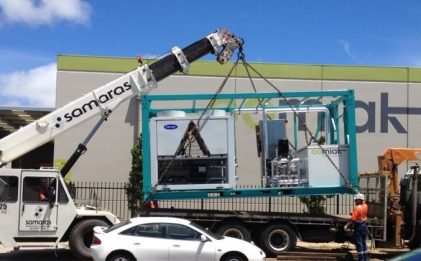 a crane lifting a containerized chilled water supply rig onto a truck with a white car in front of it