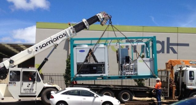 a crane lifting a containerized chilled water supply rig onto a truck with a white car in front of it